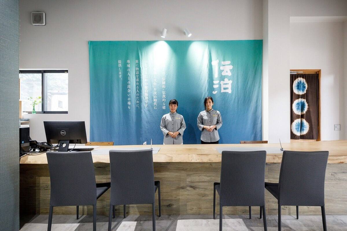 A modern conference room with a long table, four chairs, and two individuals standing in front of a backdrop with Japanese text.