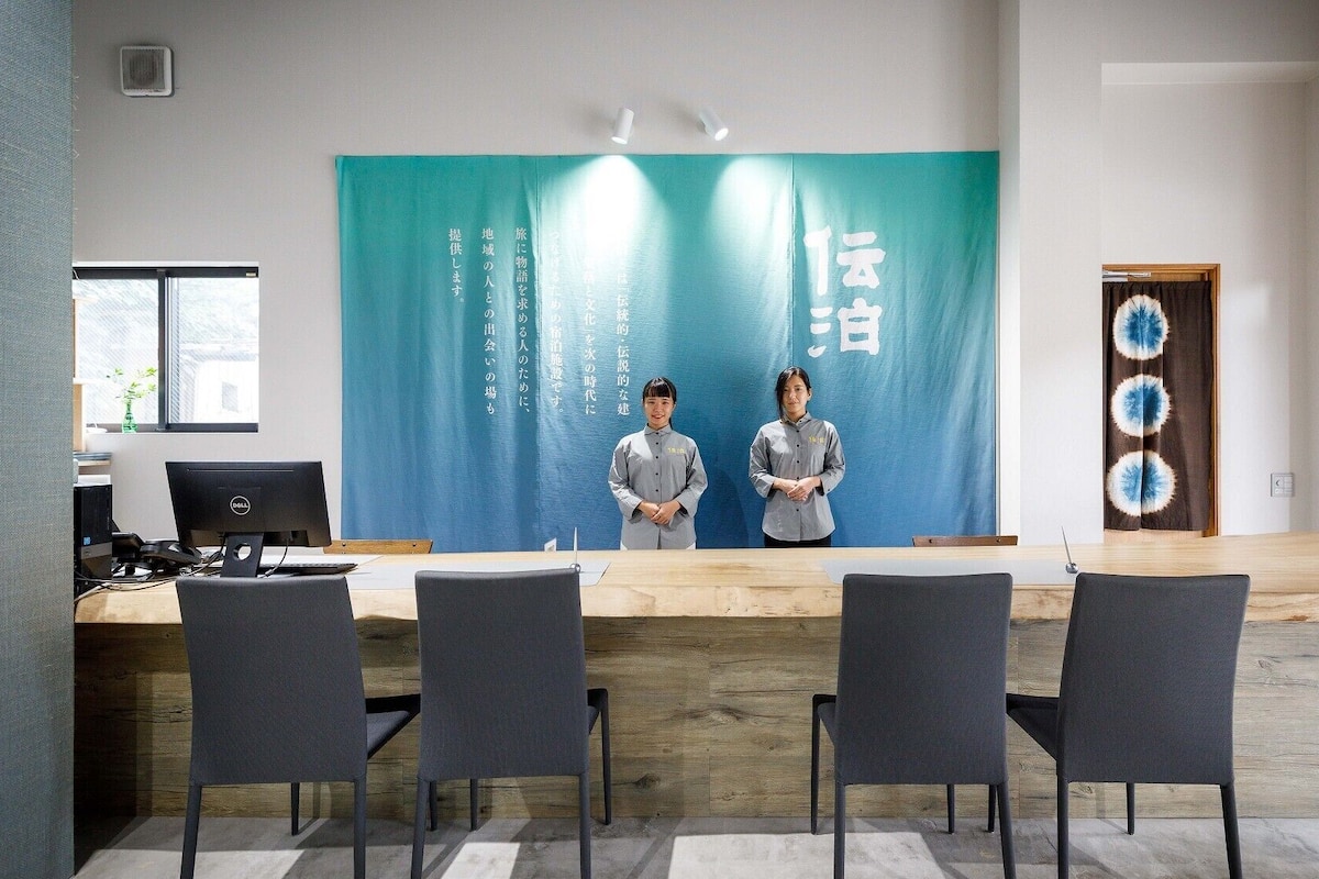 Moderni kokoushuone, jossa on pitkä pöytä, neljä tuolia, tietokone ja sininen taustaseinä, jossa on japaninkielistä tekstiä.