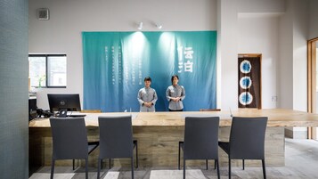 Un salón de conferencias moderno con una larga mesa, cuatro sillas y dos personas de pie frente a un fondo con texto japonés.