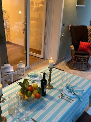A dining area with a table set for two, featuring a striped tablecloth, a candle, a bottle of wine, and a fruit bowl.