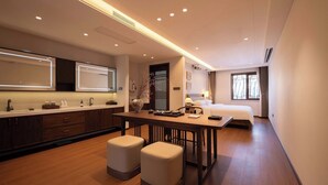Quarto de hotel moderno com piso de madeira, área de refeições com mesa e cadeiras, cama, espelho grande e janela.
