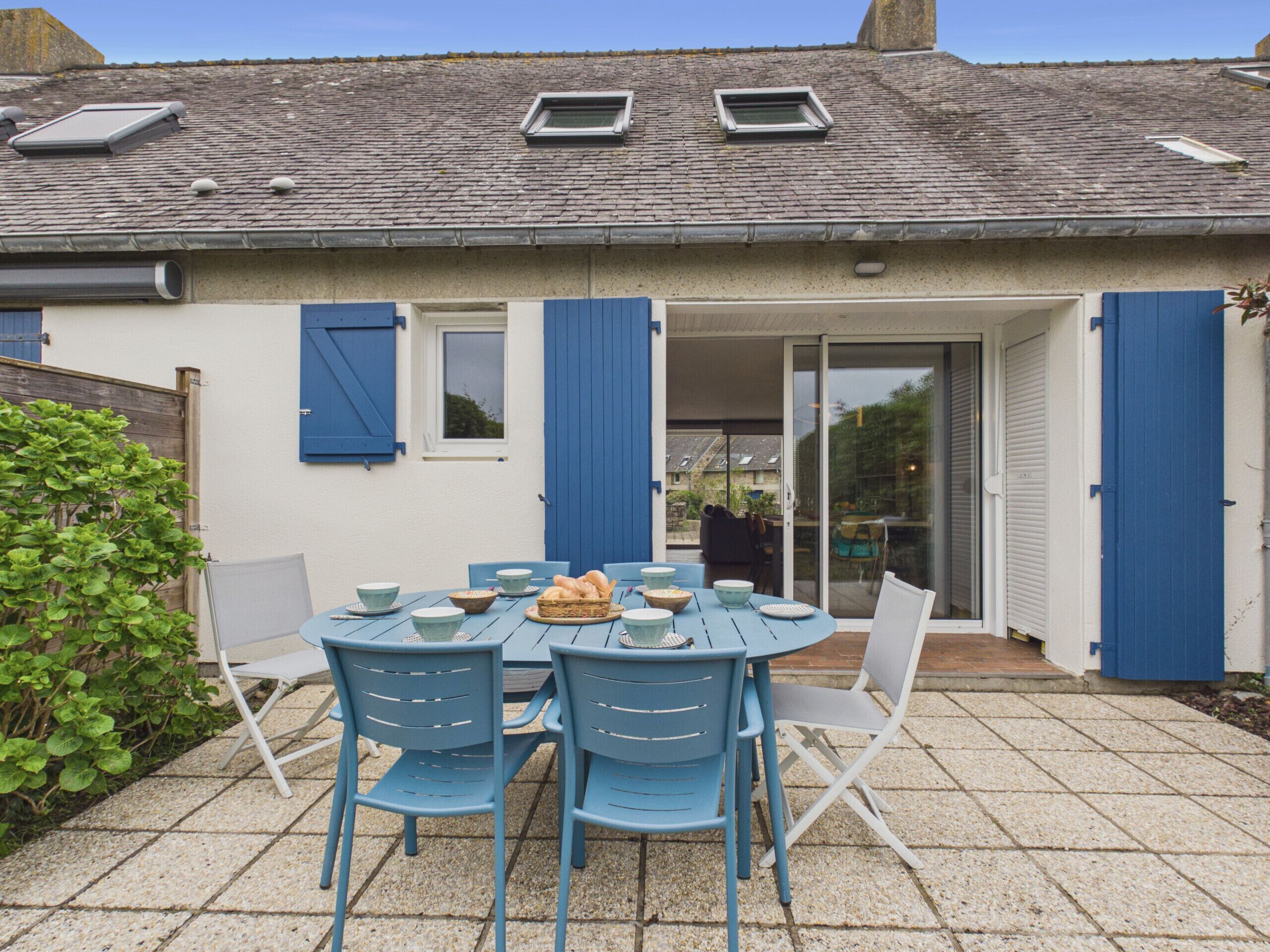 A patio with a table and chairs, a small garden, and a building with blue shutters and a tiled roof.