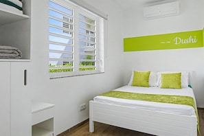A hotel room with a bed, a window with blinds, a wall-mounted air conditioner, and a sign with the name 'Dushi'. - Luxury New 2BR Apartment near Blue Bay Beach, 20m Pool, Sleeps max 4 (Sint Michiel)