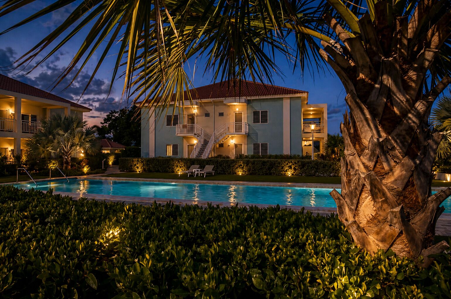 A hotel with a swimming pool, palm trees, and a well-lit exterior at dusk.