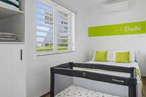 A room with a crib, a window with blinds, a wall-mounted air conditioning unit, and a bed with pillows. - Luxury New 2BR Apartment near Blue Bay Beach, 20m Pool, Sleeps max 4 (Sint Michiel)