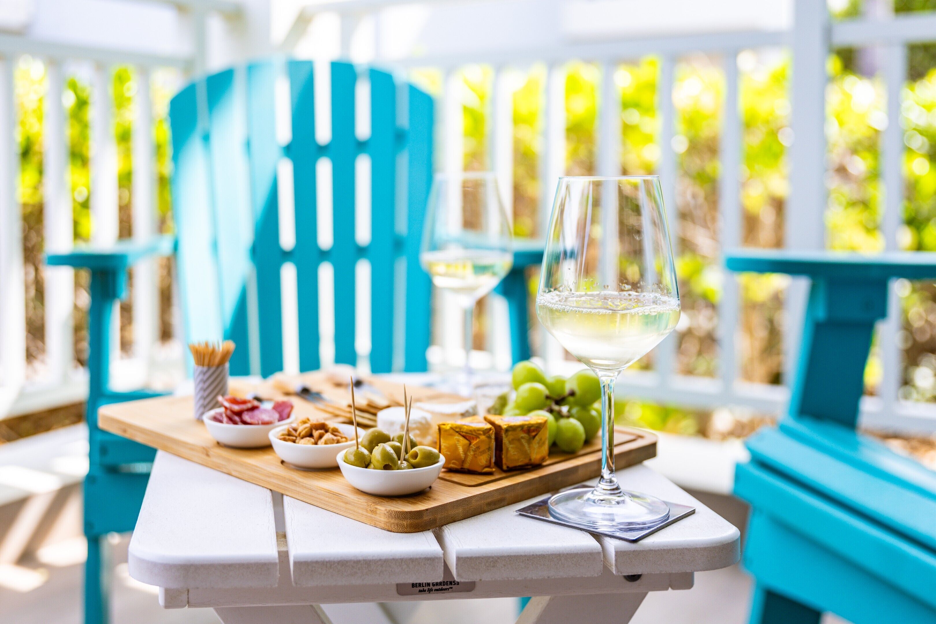 A table with food items and a glass of white wine on a terrace with blue chairs and a white fence in the background.