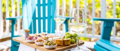 Ein Tisch mit Speisen und einem Glas Weißwein auf einer Terrasse mit blauen Stühlen und einem weißen Zaun im Hintergrund.