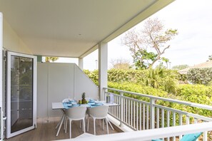 A balcony with a table set for two, white wicker chairs, and a view of a garden with trees and shrubs. - Luxury New 2BR Apartment near Blue Bay Beach, 20m Pool, Sleeps max 4 (Sint Michiel)