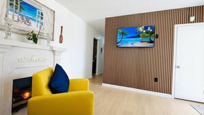 A living room with a yellow sofa, a fireplace, a TV mounted on a wooden wall, and a door. - Home Near Hard Rock w/ BBQ & Pool Table (Opa-locka)