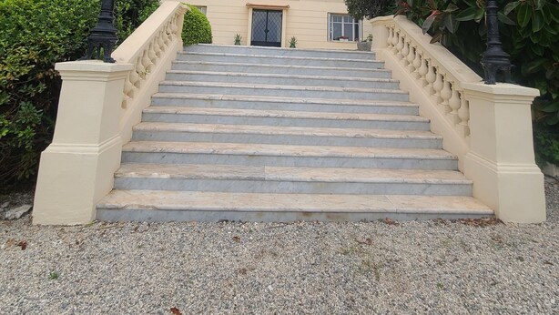 Un immeuble à plusieurs étages, doté d’un grand escalier menant à un balcon, entouré d’une végétation luxuriante et avec des montagnes en arrière-plan.