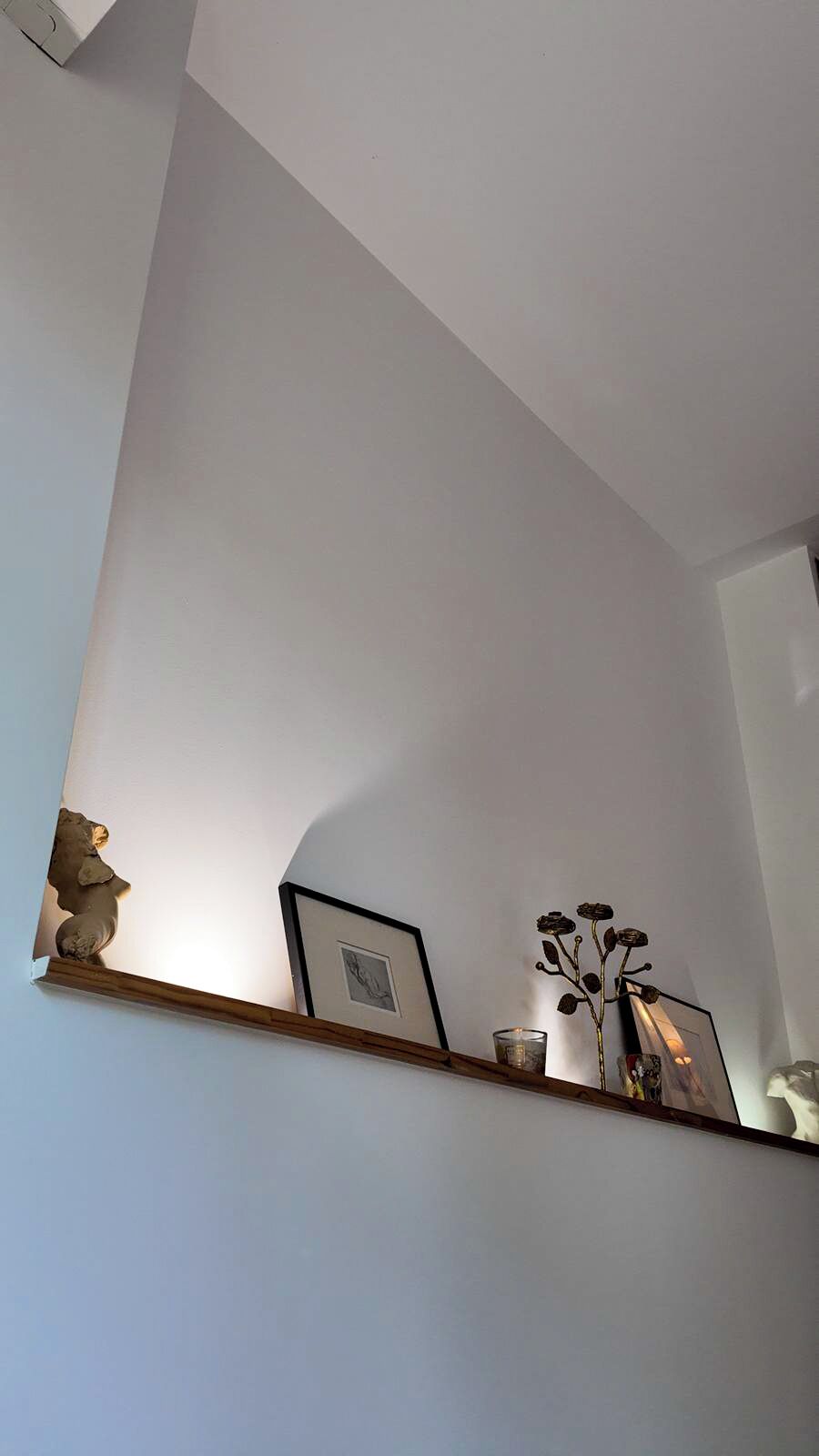 A room with a white wall, a wooden shelf with a framed picture, a small vase with dried flowers, and a candle.