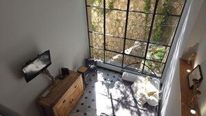 Une pièce avec une grande fenêtre, une commode en bois, un petit tabouret, un lit recouvert d’une couverture blanche et un téléviseur fixé au mur.