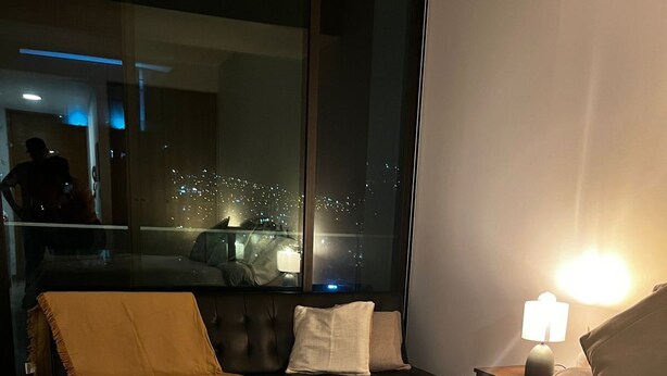 Quarto de hotel com uma cama grande, vista da cidade à noite, varanda e mesa de cabeceira com abajur.