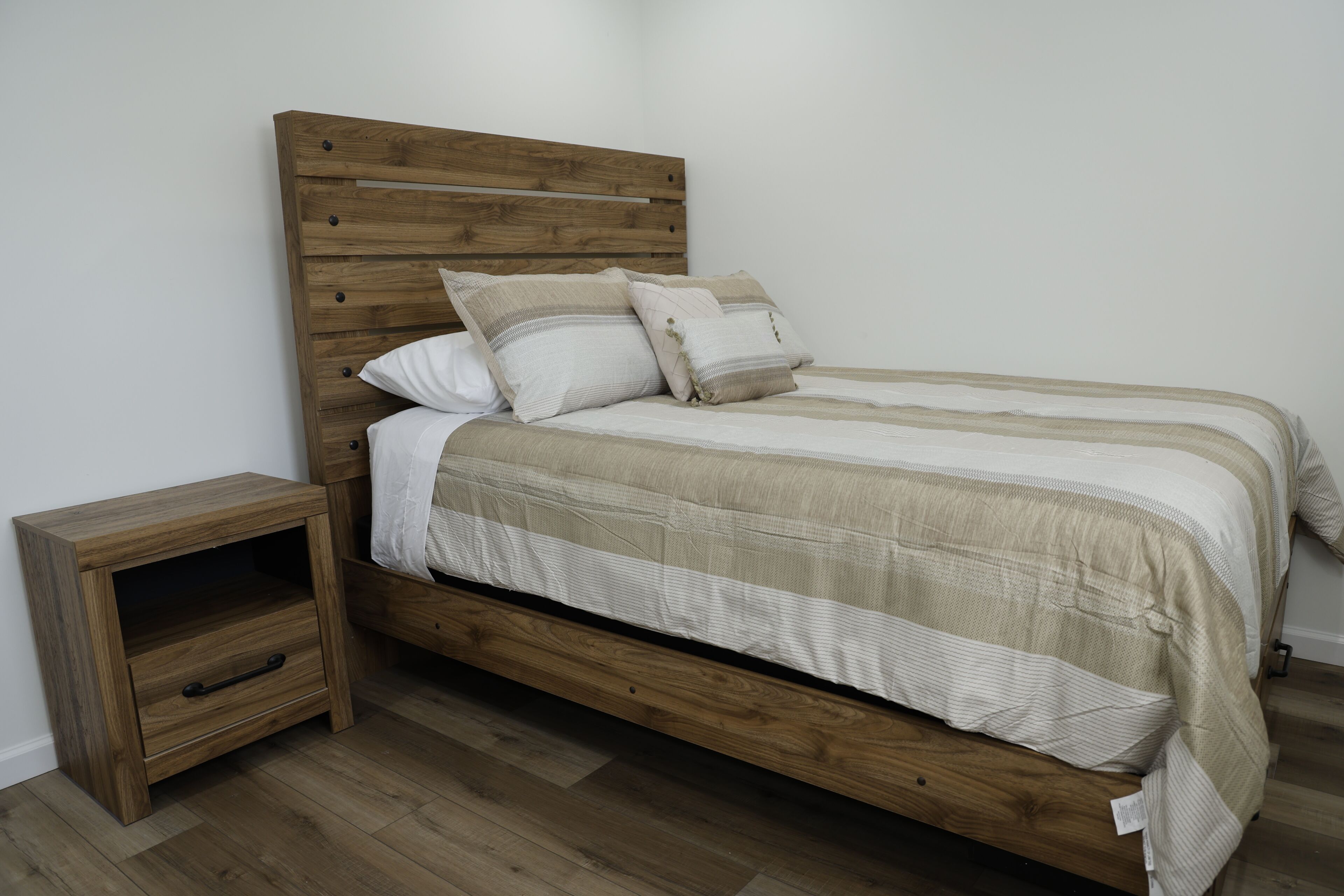 A wooden bed with a headboard, two pillows, a nightstand with a drawer, and a striped bedspread.