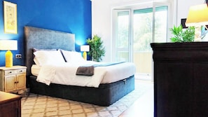 A bedroom with a large bed, a nightstand with a lamp, a dresser, and a view of the outdoors through a glass door.