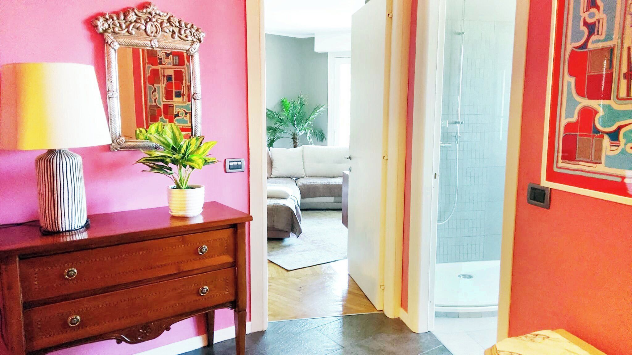 Una habitación con un aparador de madera, un espejo, una planta en maceta y una puerta que da a un baño con ducha.