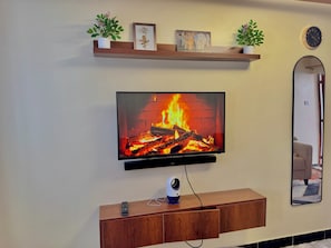 A flat-screen TV mounted on a wall with a fireplace below it, a wooden console table with a remote control, and a mirror on the adjacent wall.