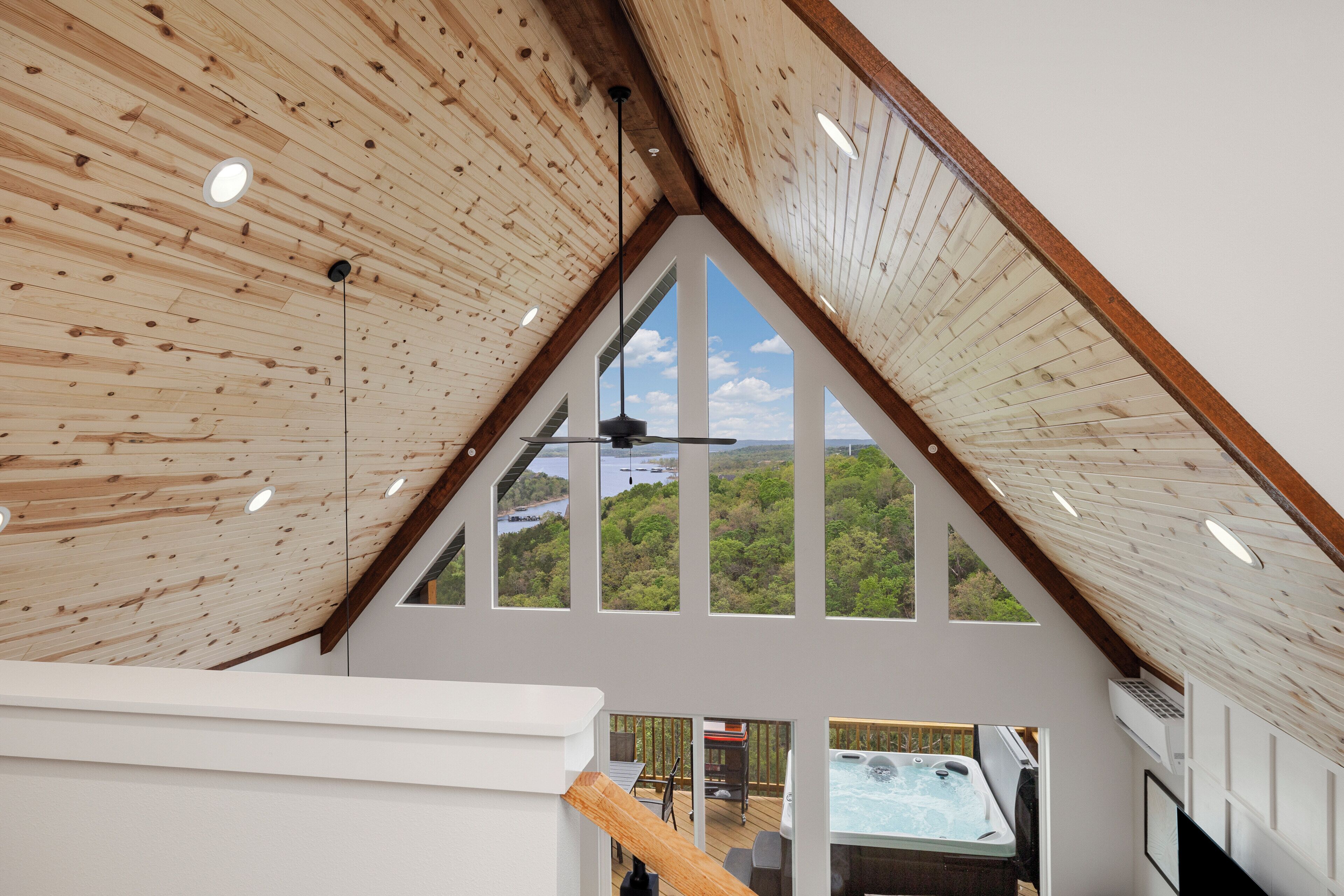 A high-ceilinged room with wooden beams, large windows, and a view of a hot tub outside.