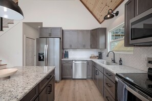 A modern kitchen with a stainless steel refrigerator, dishwasher, and oven, wooden flooring, and a window with a view. - Modern A-Frame + Hot Tub with Lake Views Near Branson (MARVEL CAV PK)