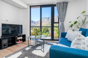 A modern living room with a large flat-screen TV, a blue sofa, a small table, and a view of mountains through large windows. - The Cedar 202 (Cape Town)