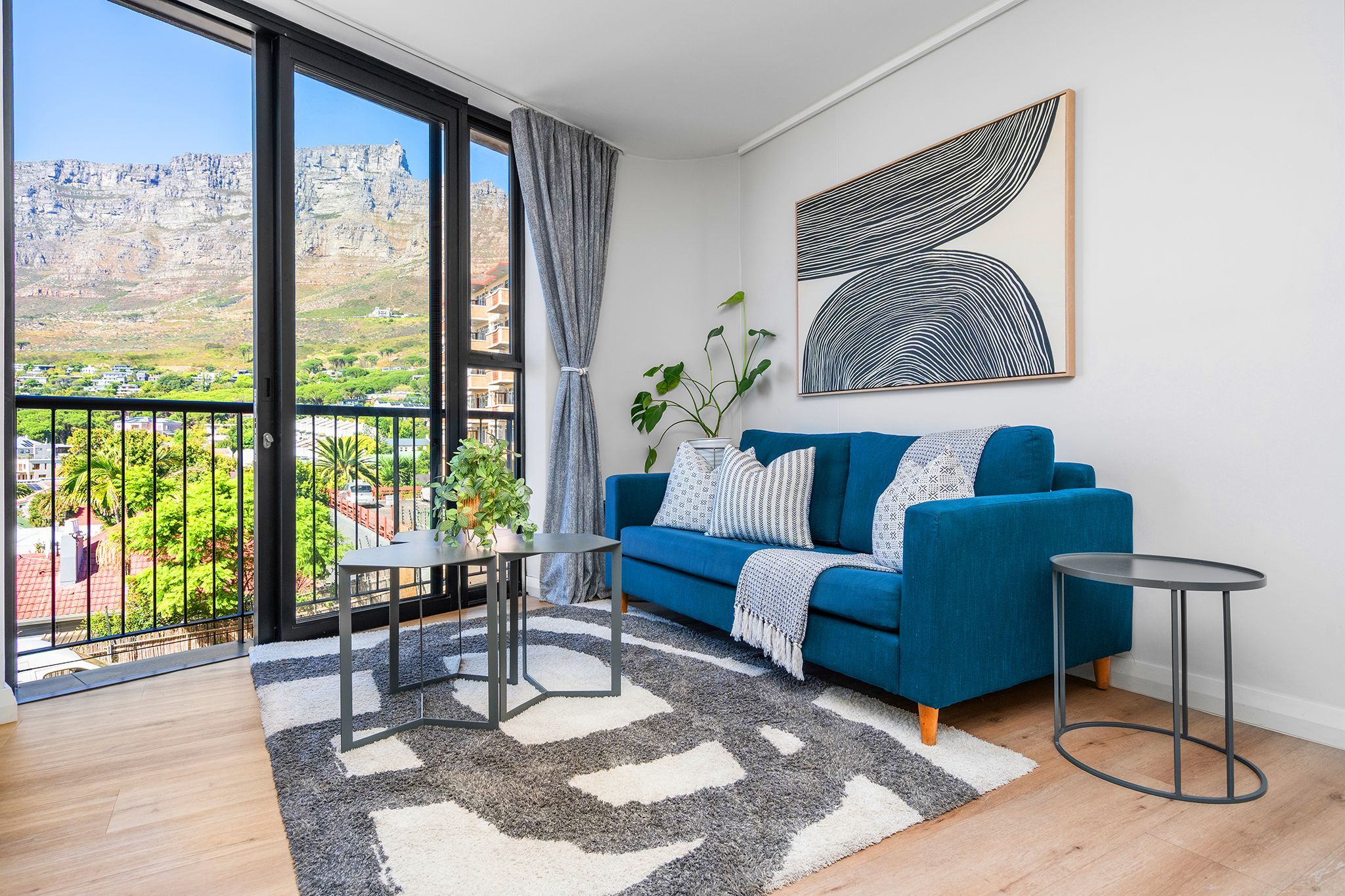 A living room with a blue sofa, a round table, a large window with a view of a mountainous landscape, and a modern abstract painting on the wall.