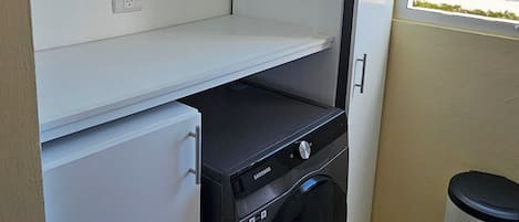 Uma área de lavanderia compacta, com máquina de lavar, pequeno frigorífico e um cesto de lixo.