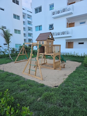 Parquinho infantil com balanço de madeira e estrutura de escalada, cercado por área arenosa e vegetação, em frente a um edifício moderno branco com várias janelas e varandas.