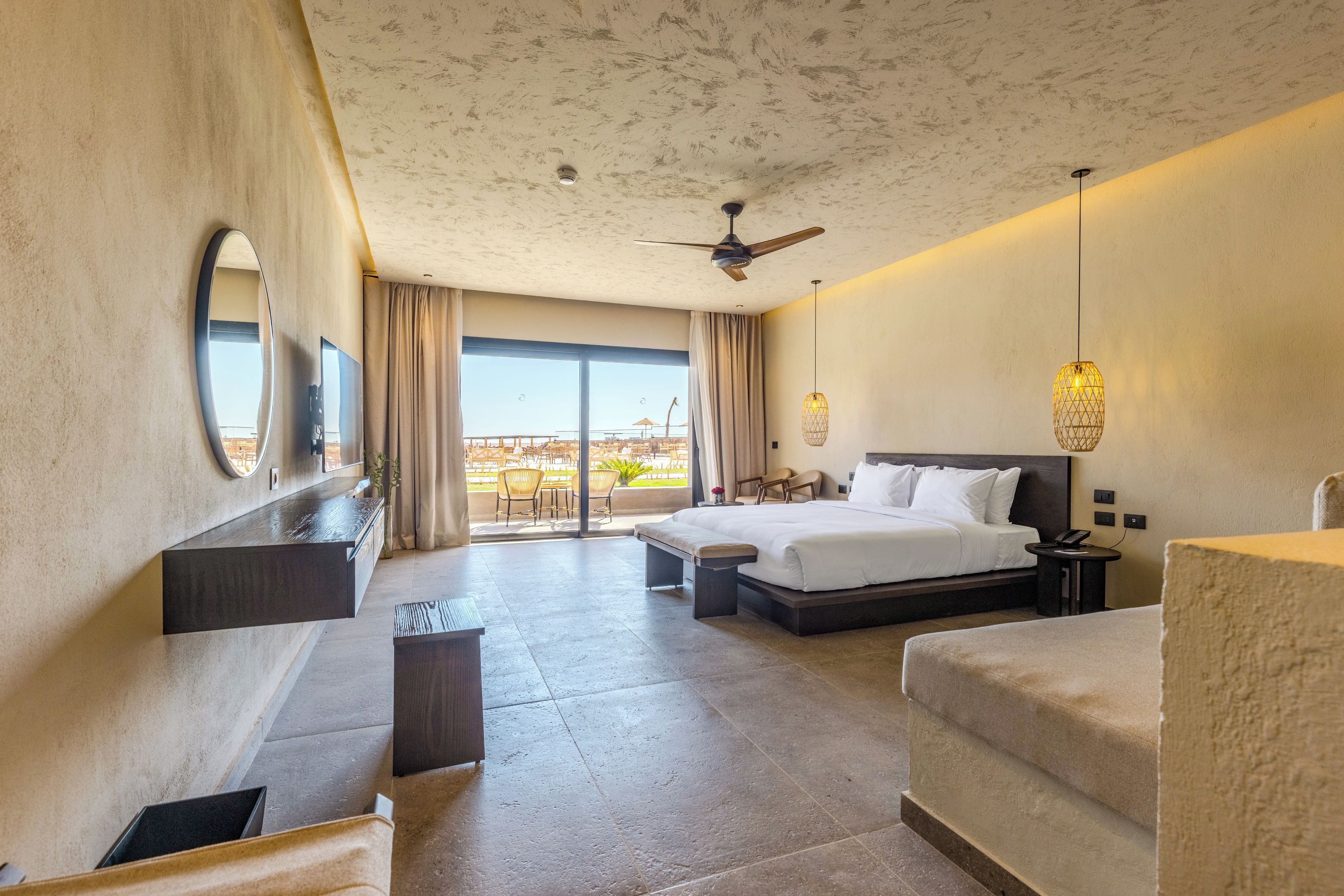 Habitación de hotel con cama, escritorio, silla, balcón con mobiliario exterior y ventilador de techo.