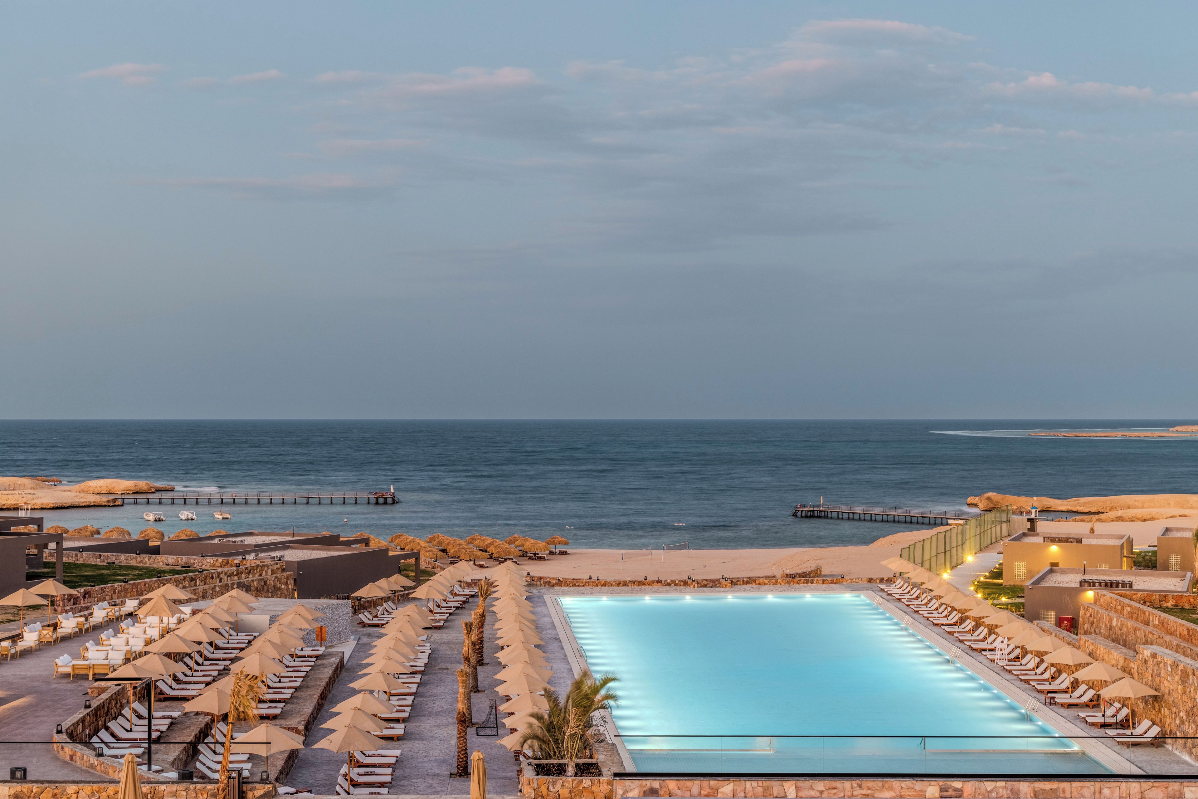 A resort with a swimming pool, beach umbrellas, lounge chairs, and a view of the sea.