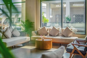 Um saguão de hotel moderno com área de estar, incluindo sofás e poltronas, uma mesa de centro de madeira e janelas amplas com vista para a paisagem urbana.