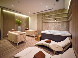 Um quarto de spa com mesa de massagem, cadeira de massagem, hidromassagem e uma pequena mesa com uma vela.