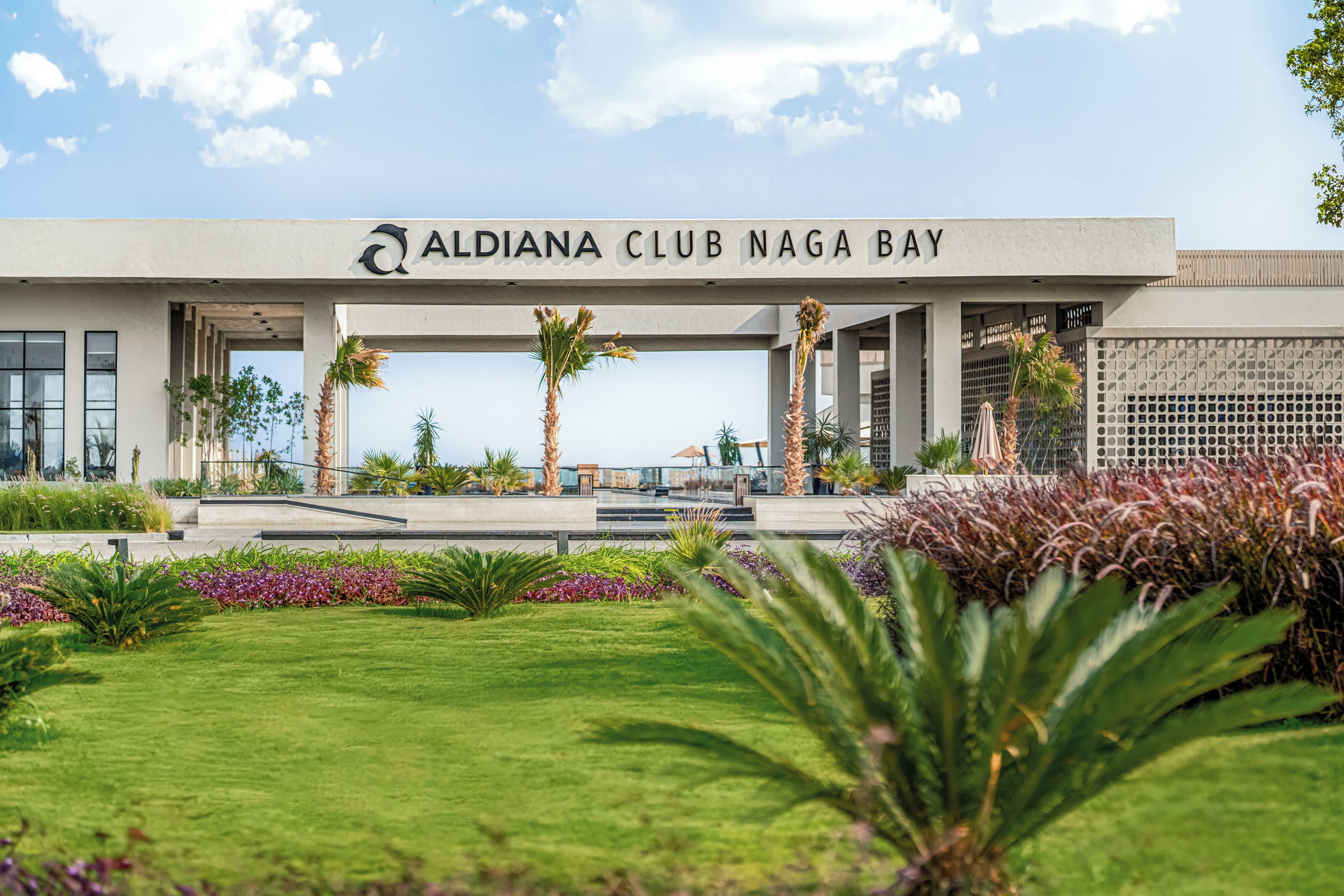 The exterior of Aldiana Club Naga Bay with a clear sky, palm trees, and well-maintained landscaping.
