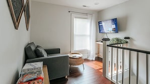 Uma sala de estar moderna com um sofá cinzento, uma mesa redonda pequena e uma TV fixada na parede.