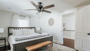 Quarto com cama, um banquinho, ventilador de teto, mesa de cabeceira, abajur, janela com cortinas, um objeto decorativo na parede e porta para outro cômodo.