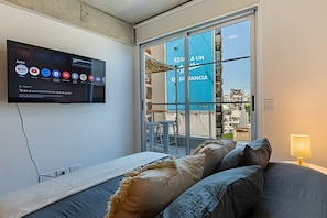 Ein Hotelzimmer mit Bett, Wandfernseher und Blick auf eine Stadtlandschaft durch große Fenster.