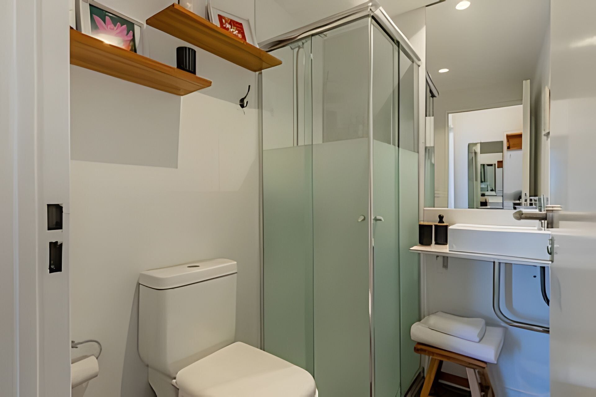 Un baño moderno con inodoro, cabina de ducha de vidrio, lavamanos con espejo y estantes de madera sobre el inodoro.