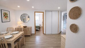 A room with a dining table set for four, a bed with a green bedspread, a wooden door, and a wall with woven wall art. - LVAM - Renovated 2-bedroom apartment – Beach – Garden – Golf 5 (Agde)
