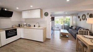 A modern kitchen with white cabinets, a black oven, and a coffee maker. There is a dining area with a table and chairs, a sofa, and a lamp. The room has a large window and a view of the outdoors. - LVAM - Renovated 2-bedroom apartment – Beach – Garden – Golf 5 (Agde)