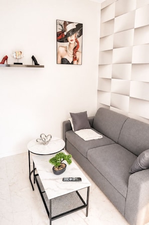 Een moderne woonkamer met een grijze bank, een witte salontafel met een plant, een ingelijst schilderij aan de muur, en een paar hoge hakken op een plank.