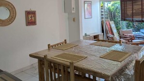 Un comedor con una mesa cubierta de plástico, sillas de madera, un ventilador de techo y vistas al exterior a través de una ventana.