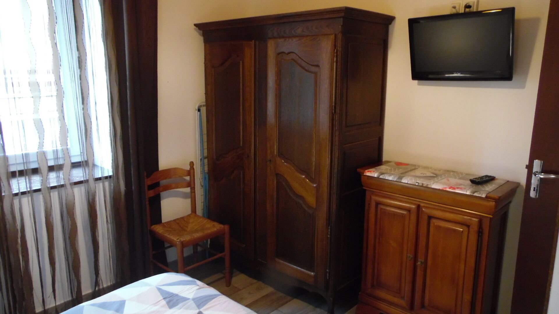 Um quarto com cama, guarda-roupa de madeira, cadeira, televisão e janela com cortinas.