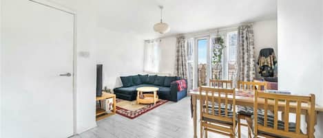 A living room with a sofa, a dining table with chairs, a TV, and a window with curtains.