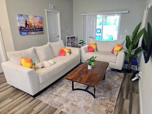 A living room with two white sofas, colorful cushions, a wooden coffee table, and a cityscape painting on the wall. - Family Friendly Gem 10 min to Ybor & Downtown! (Tampa)