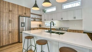 Een moderne keuken met houten kastjes, een groot eiland, barkrukken, hanglampen en een roestvrijstalen koelkast.