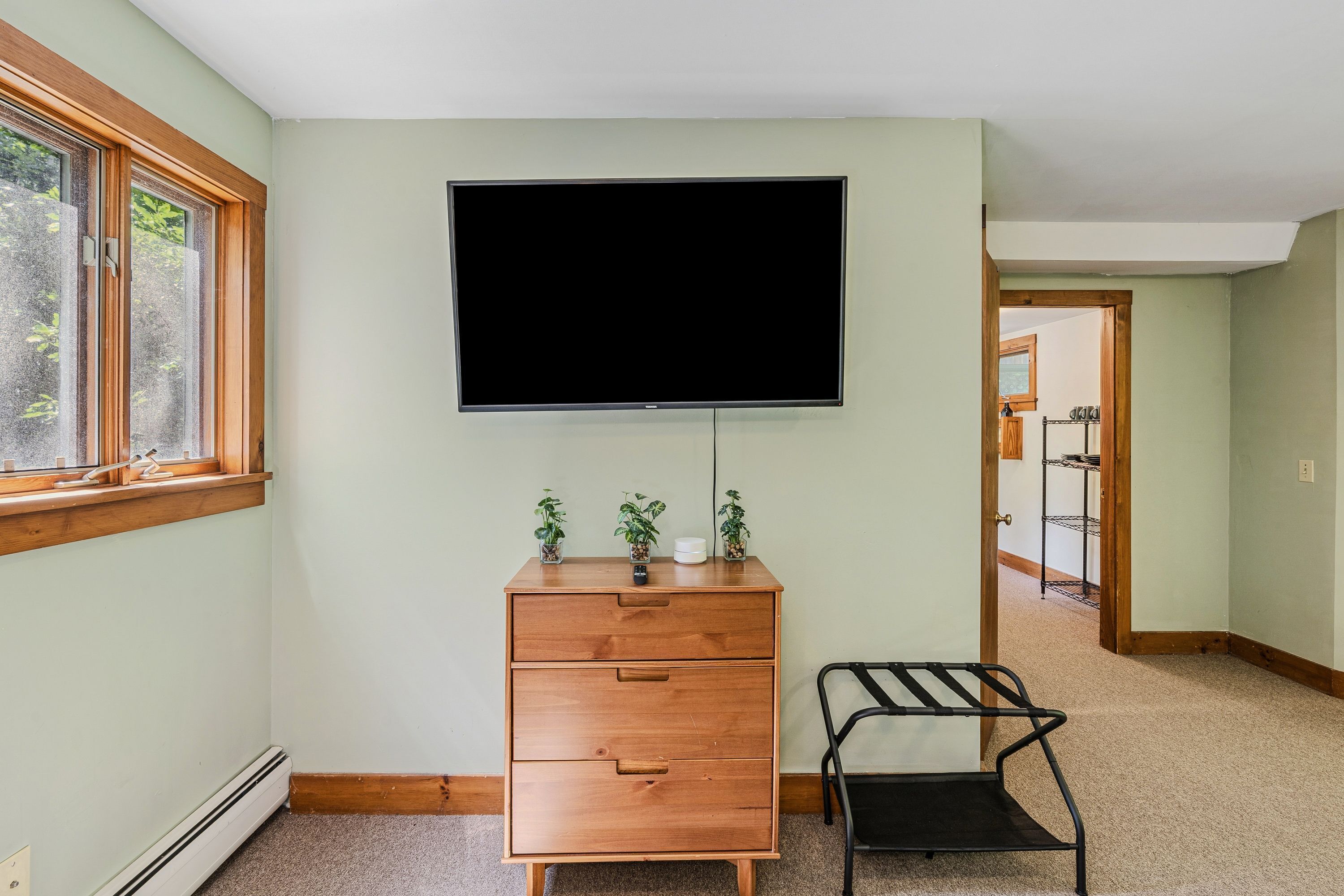 A room with a wooden dresser, a television mounted on the wall, a window with blinds, and a door leading to another room.