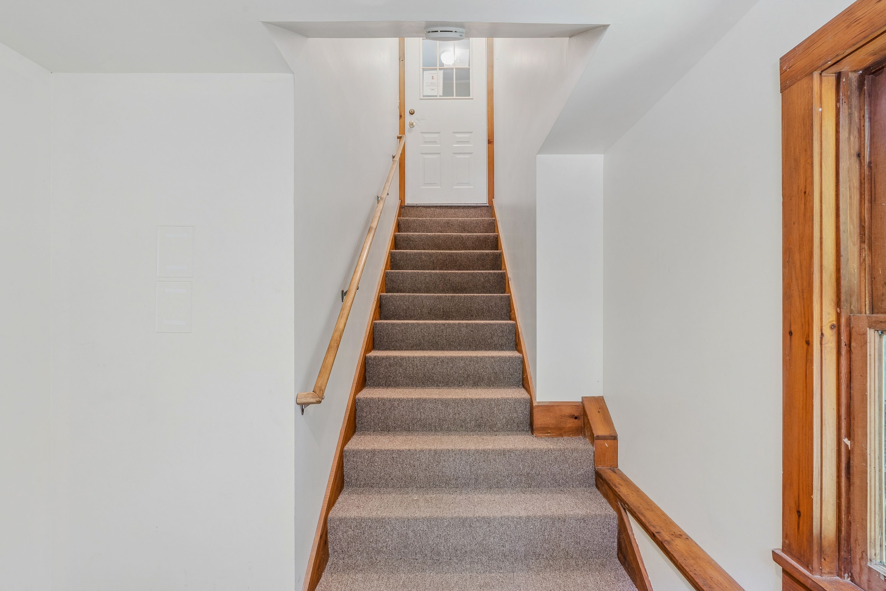 Un escalier dont les marches sont recouvertes de moquette et qui possède des rampes en bois, menant à une porte blanche surmontée d’une petite fenêtre.