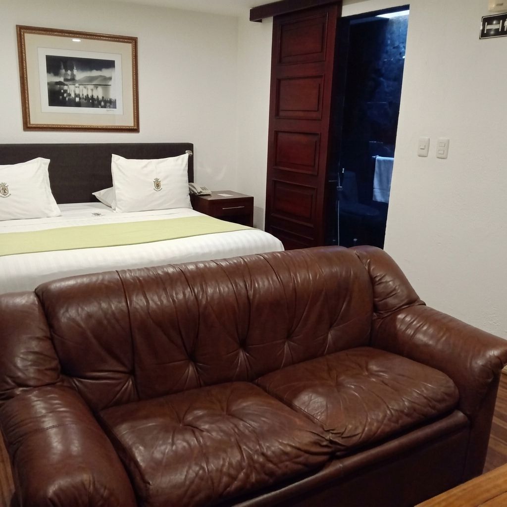 Une chambre d’hôtel avec un lit, un canapé en cuir marron, un tableau encadré au mur et une porte en bois.