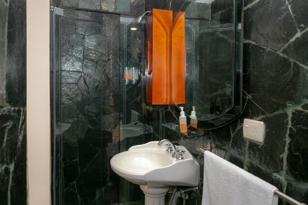 Casa de banho com lavatório pedestal branco, cabine de chuveiro em vidro e parede de mármore.