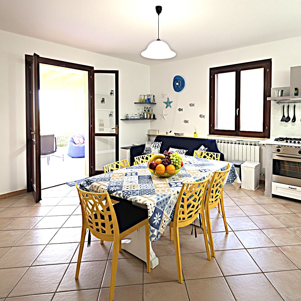 A dining area with a table set for four, a bowl of fruit, and a kitchen in the background.