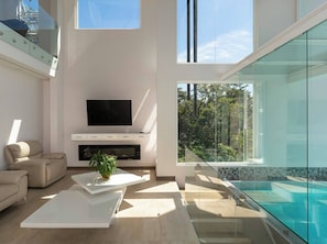 A modern living room with a flat-screen TV, a sofa, a coffee table, and a large window overlooking a pool. - Luxurious 4-bedroom House in Alajuela, Costa Rica (Guacima)